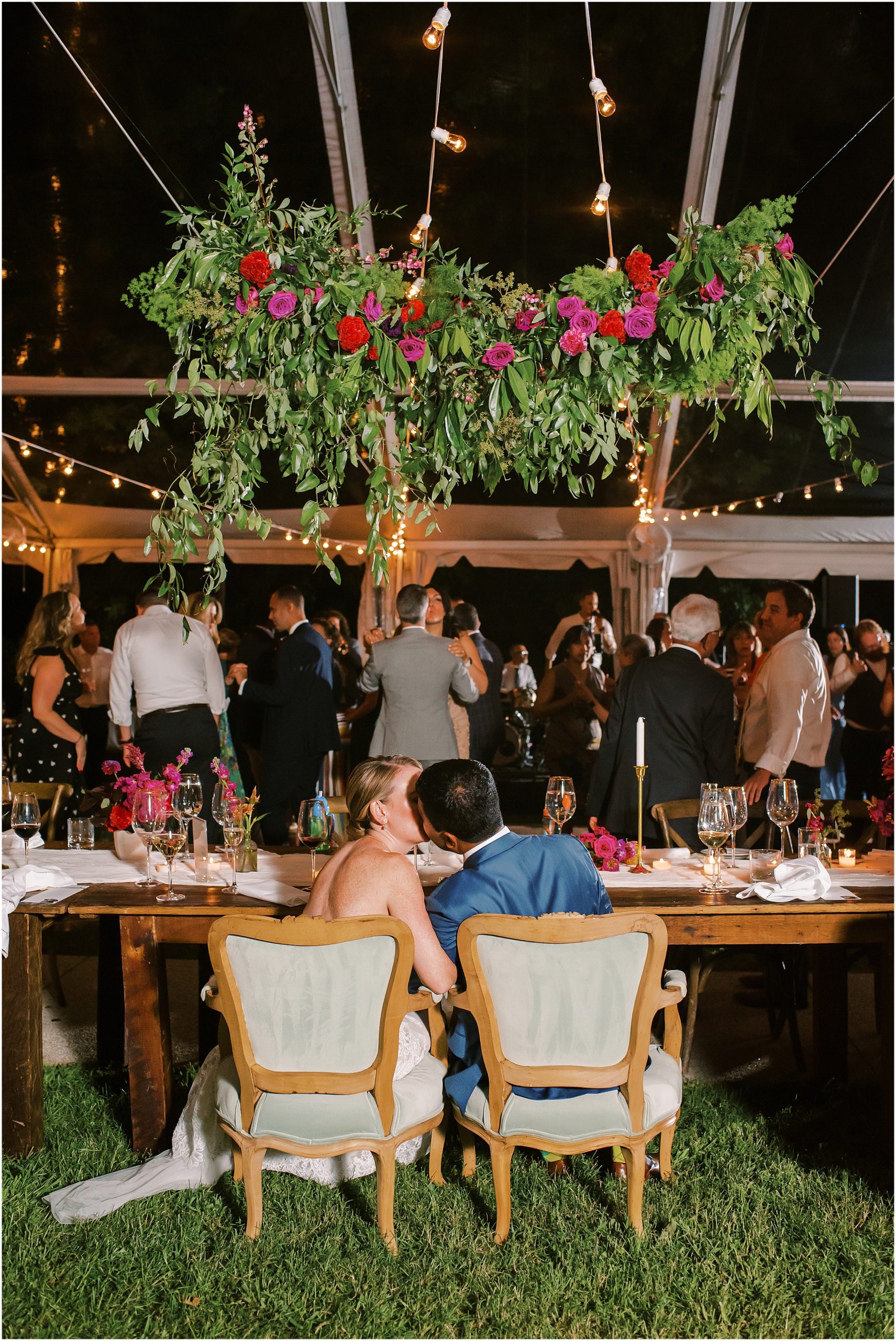 Bride and groom kissing under hanging greenery in clear wedding tent